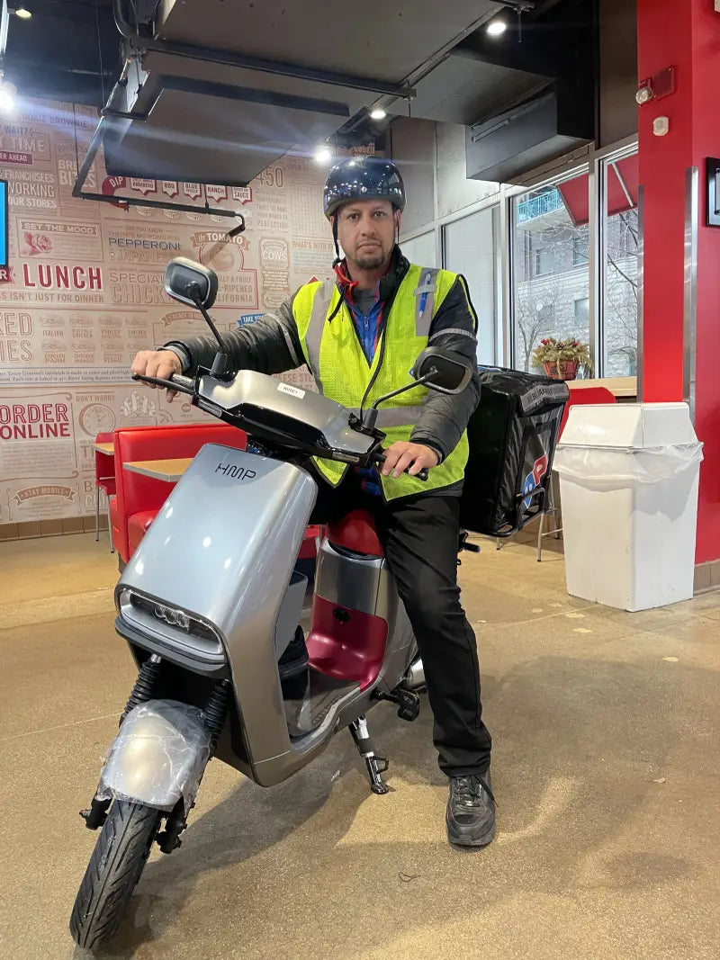 Delivery rider sitting on a silver HMP electric moped inside a restaurant, wearing a helmet and reflective safety vest.