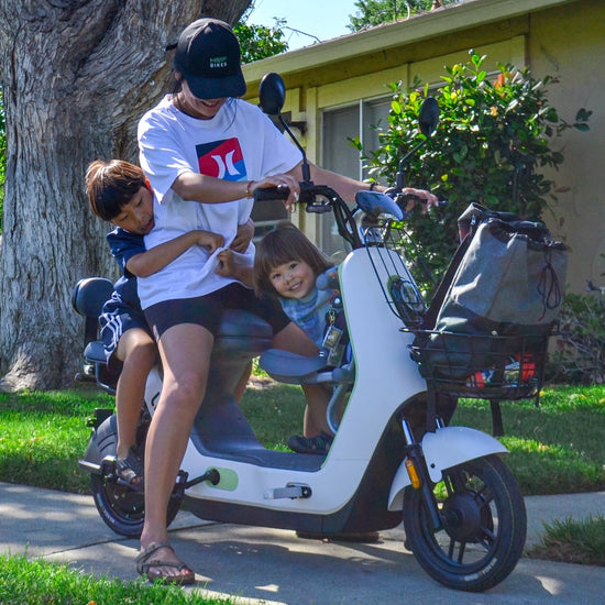 A parent riding an electric scooter with two kids on board, smiling in front of a suburban home, showcasing family-friendly design.