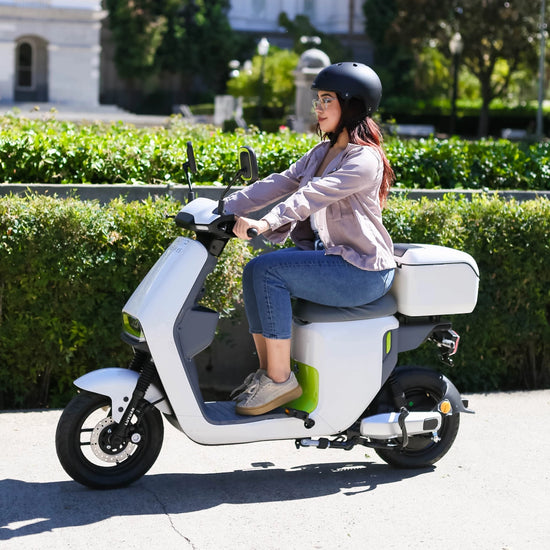 A woman riding a white electric scooter with a rear storage box, wearing a helmet and casual clothes, in a city park setting.