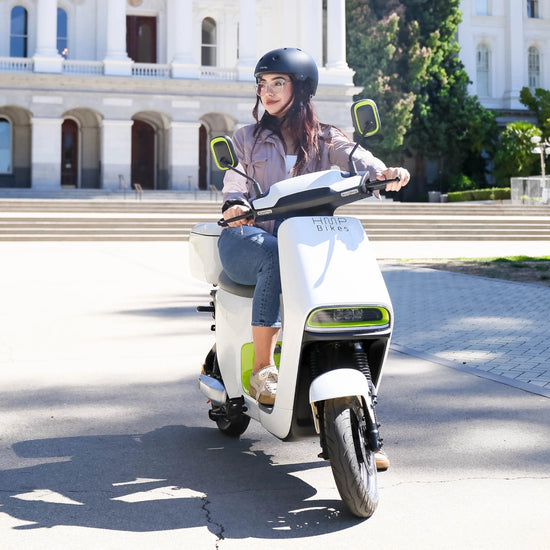 A woman riding a white HMP electric scooter in front of a historic building, wearing a helmet and sunglasses.